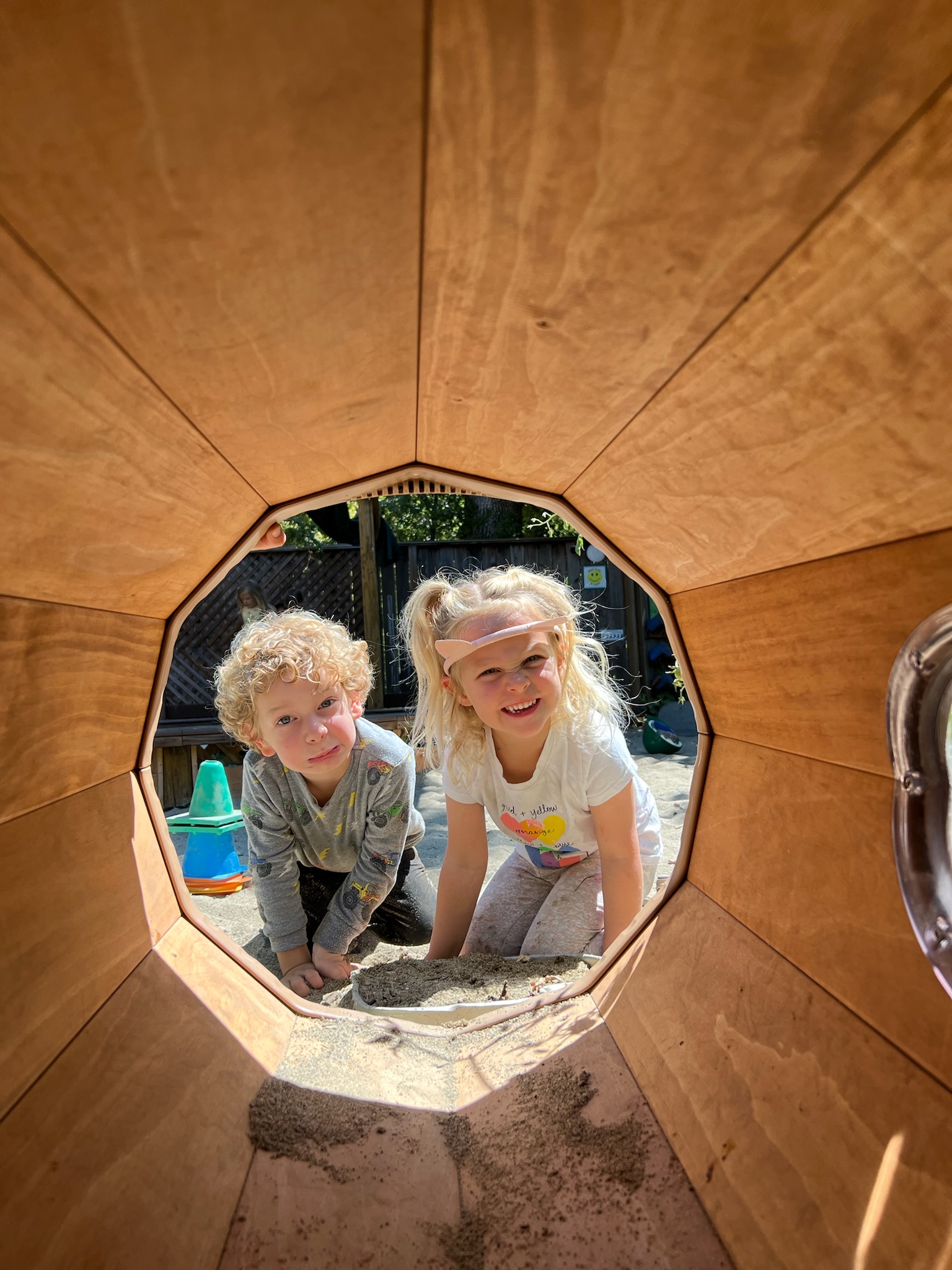Two children peeking through the wooden tunnel on the playground
